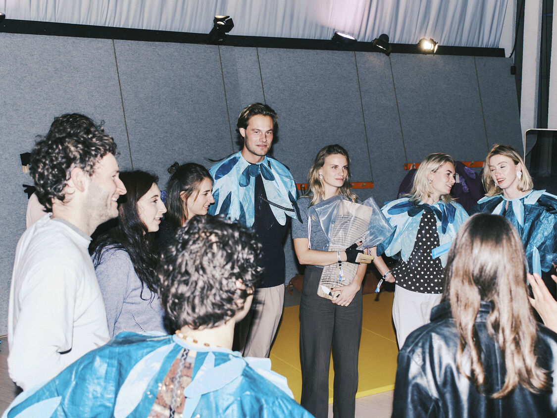 Group of people in blue leaf-shaped costumes gathered indoors; a woman in the center holds a bouquet.