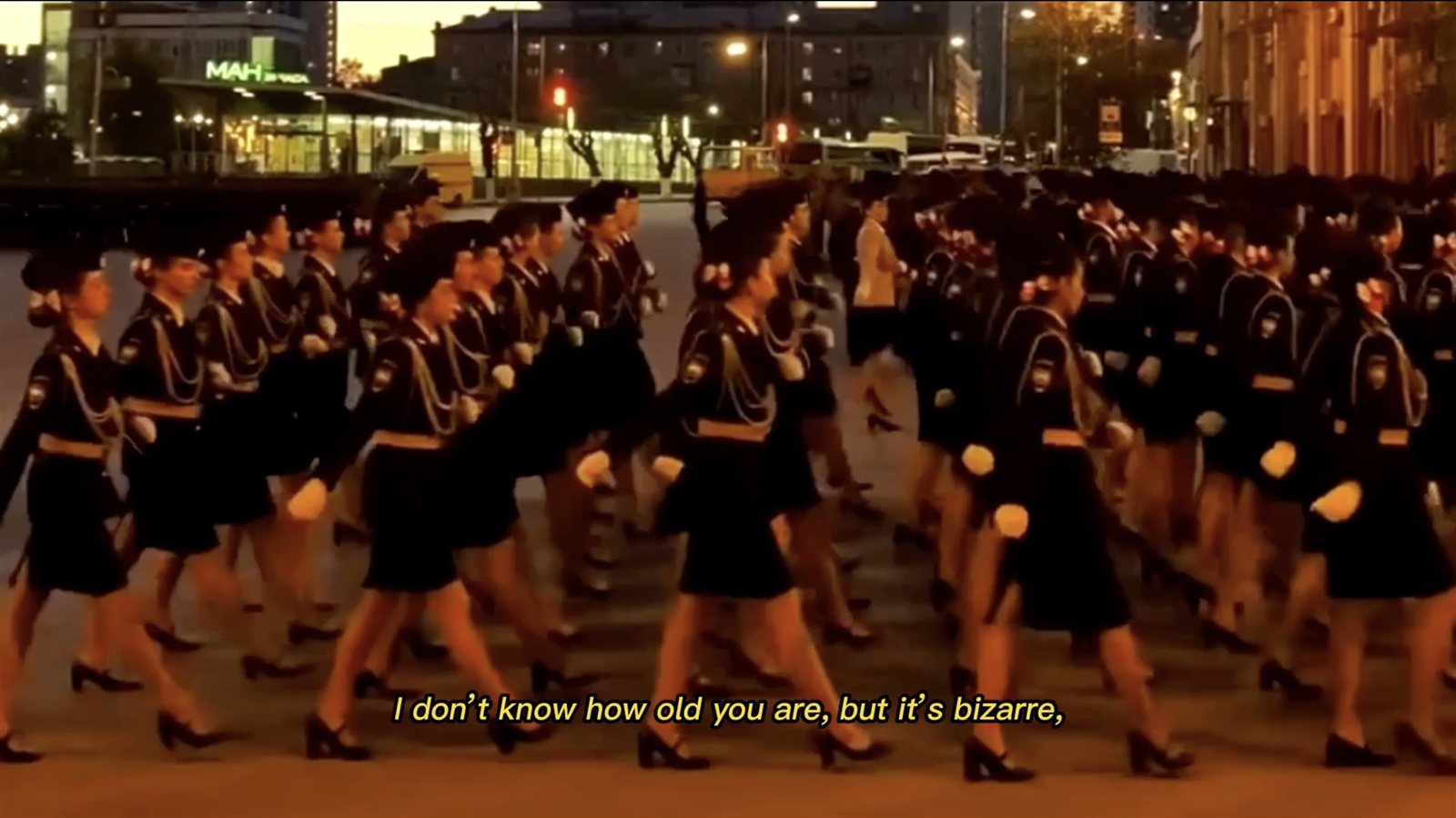 Video still of women in uniform marching through a city square at dusk, subtitled “I don't know how old you are, but it's bizarre”